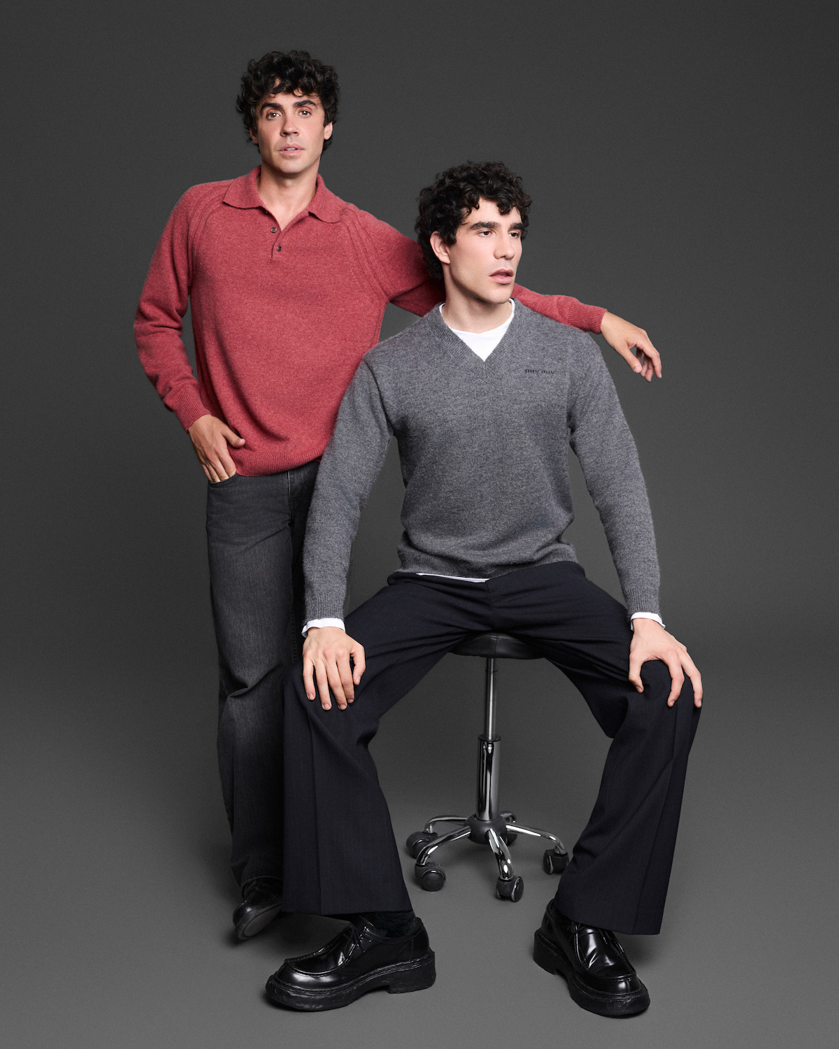 Two men pose in a studio with a dark background; one stands wearing a red sweater and jeans, while the other, dressed in a gray sweater and black pants, sits on a stool.