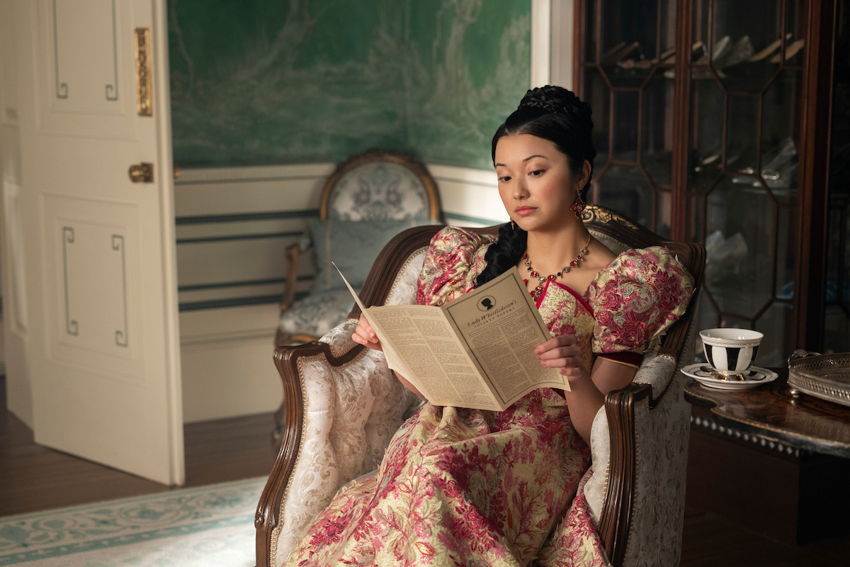 A woman in a Victorian-style dress sits in an ornate chair reading a newspaper in an elegant, vintage room with antique furniture, a teacup, and a decorative cabinet.