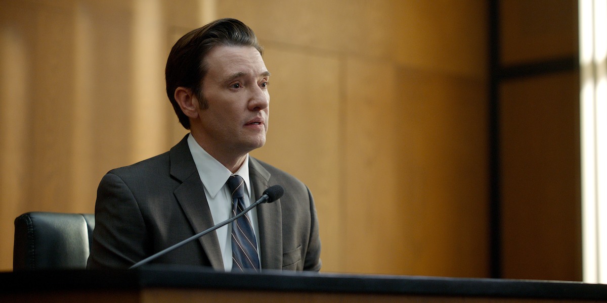 Man in a suit sitting on a witness stand, speaking into a microphone in a wood-paneled courtroom, appearing serious and attentive during a legal proceeding.