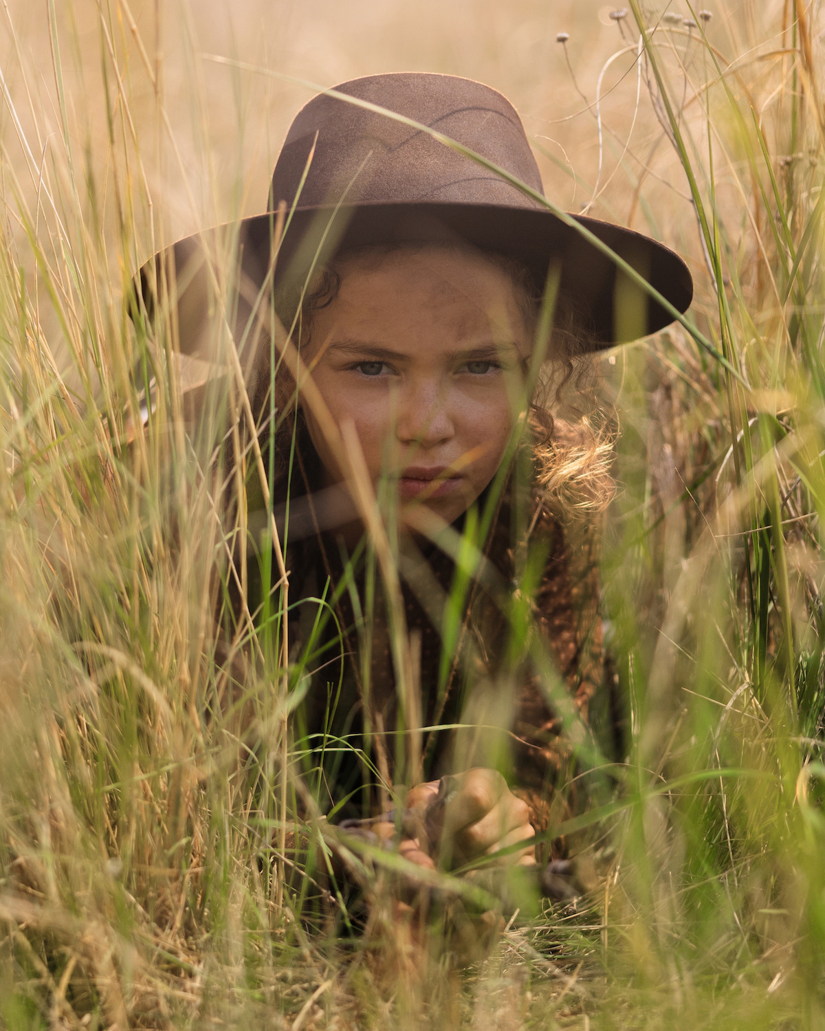 Young child wearing a wide-brimmed hat crouching in tall grass, blending into a natural outdoor setting, with sunlight filtering through the vegetation.