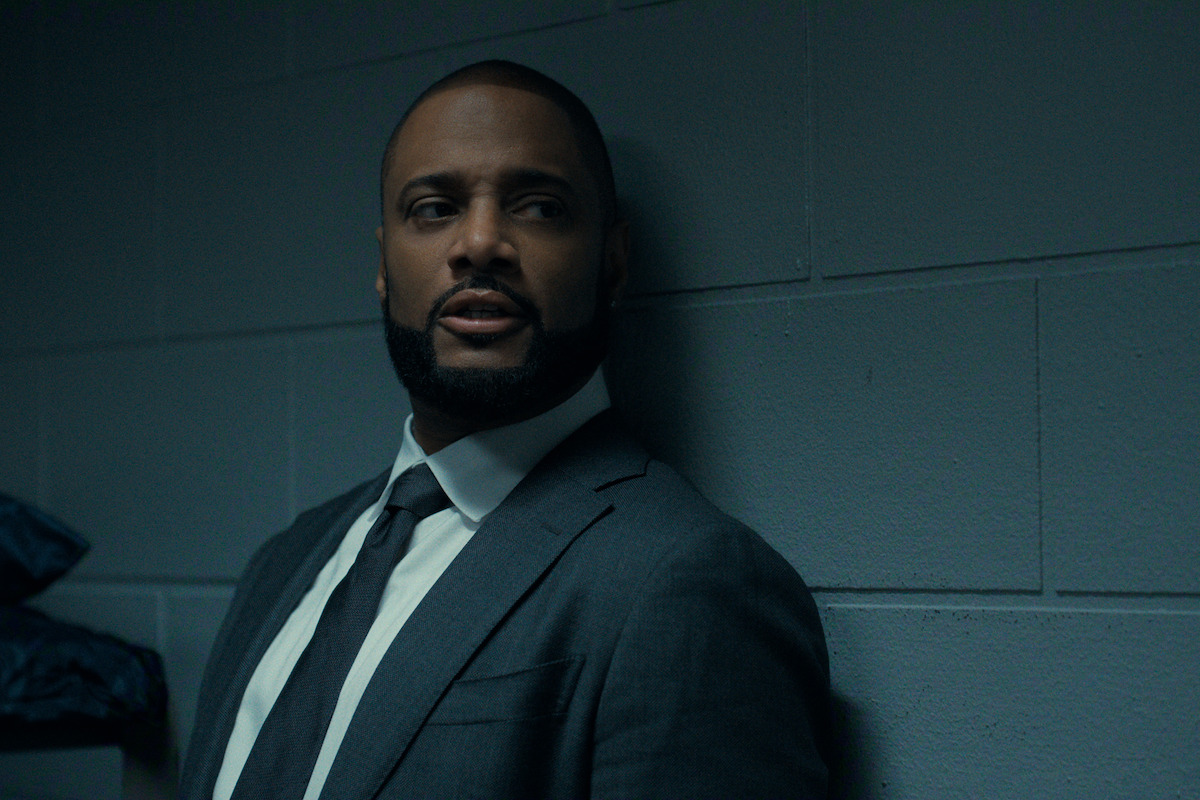 A man in a suit leans against a gray cinder block wall in a dimly lit room, looking to the side with a serious expression.
