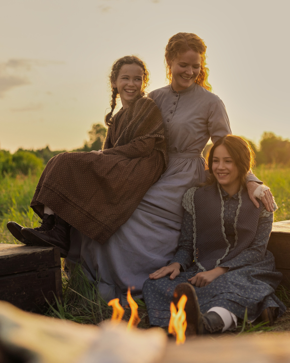 Three women in old-fashioned dresses sit outdoors on logs around a campfire at sunset, smiling and enjoying each other's company in a grassy field.