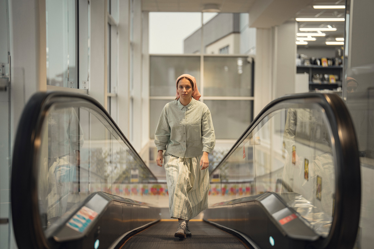 Woman in modest attire and headscarf walks up an escalator inside a modern, brightly lit building with large windows and visible store shelves in the background.