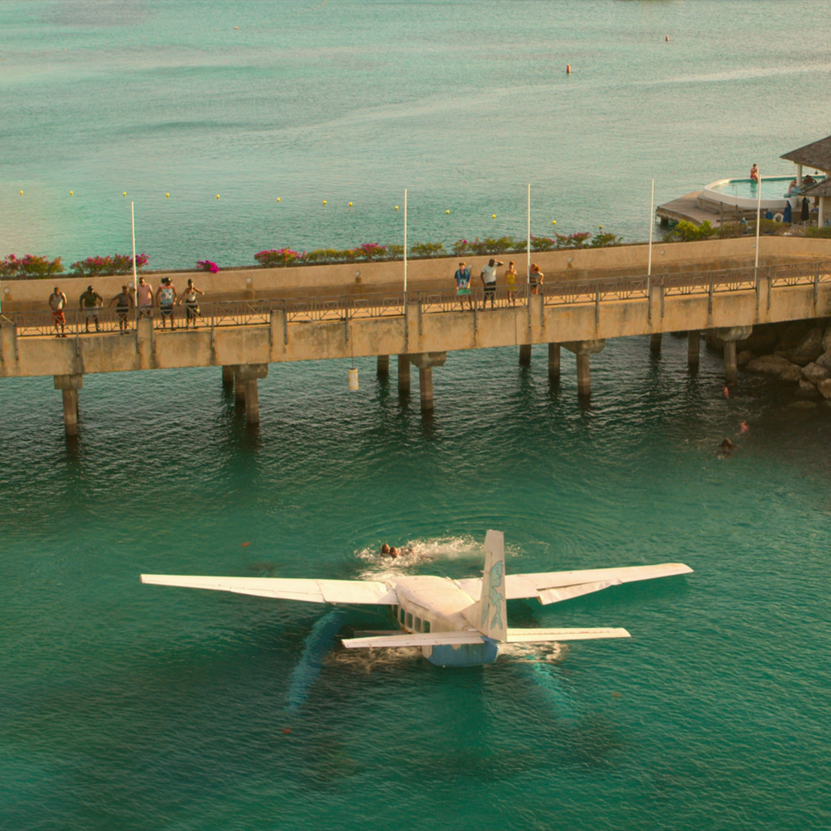 Um hidroavião flutua em águas claras perto de um píer de concreto, com várias pessoas em pé no píer e algumas visíveis na água, em um ambiente tropical à beira-mar com flores e prédios nas proximidades.