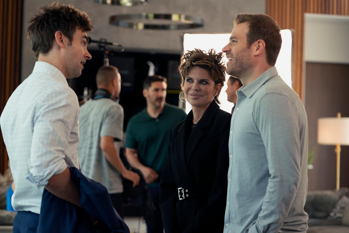 Three people have a conversation in a modern, well-lit living room with crew members, cameras, and equipment visible in the background, suggesting a film or photoshoot set environment.