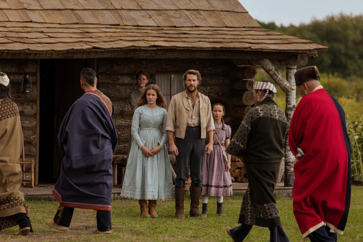 A pioneer family in historical clothing stands in front of a rustic log cabin, while several Indigenous people in traditional dress walk by, all set in a rural outdoor environment with green fields and woods in the background.