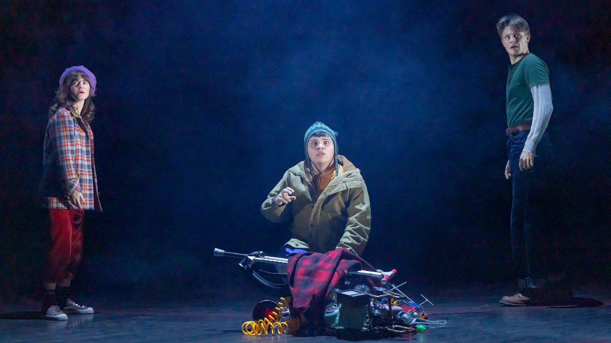 Three young people on a dimly lit stage, one kneeling beside scattered tools and props, all appearing tense or surprised; the scene has a mysterious, dramatic mood.