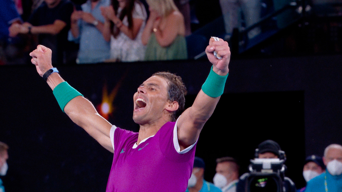 A male tennis player in a purple shirt celebrates with raised arms on a tennis court, surrounded by cheering spectators and event staff, some wearing masks, in a stadium setting.