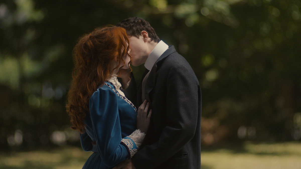 A man in a dark suit gently kisses a red-haired woman in a blue vintage dress on the forehead while standing in a sunlit, green outdoor garden or park.