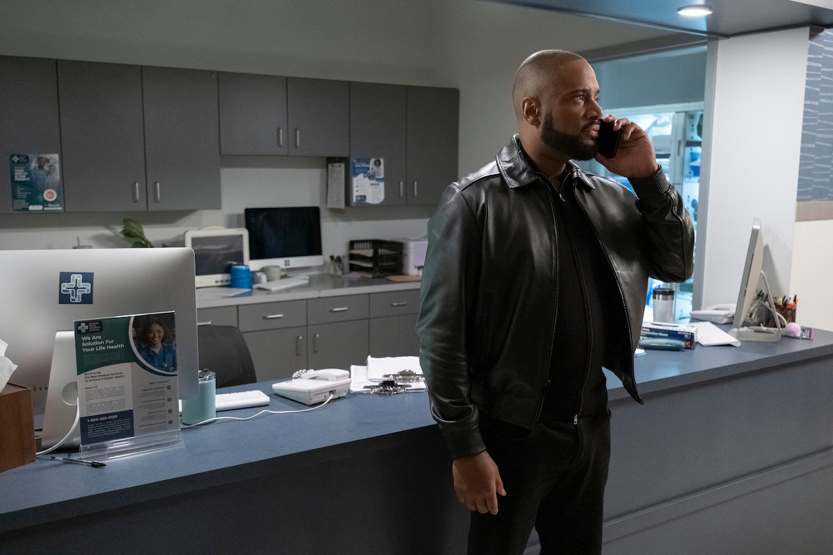 Man in black leather jacket stands at a modern medical office reception desk, talking on his phone. The environment is clean and organized with office supplies, a computer, and cabinets in the background.