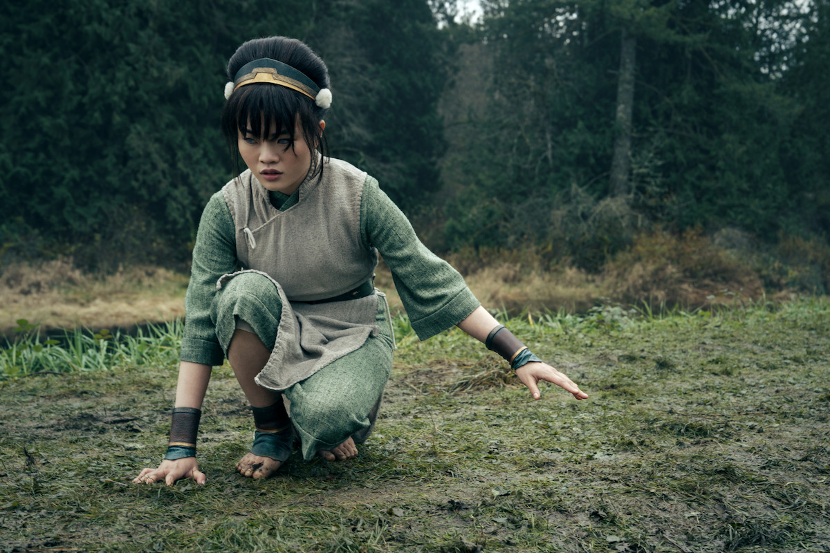 Miyako as Toph with black hair wearing green clothes crouches down in the woods with bare feet on grass and right hand touching the ground.
