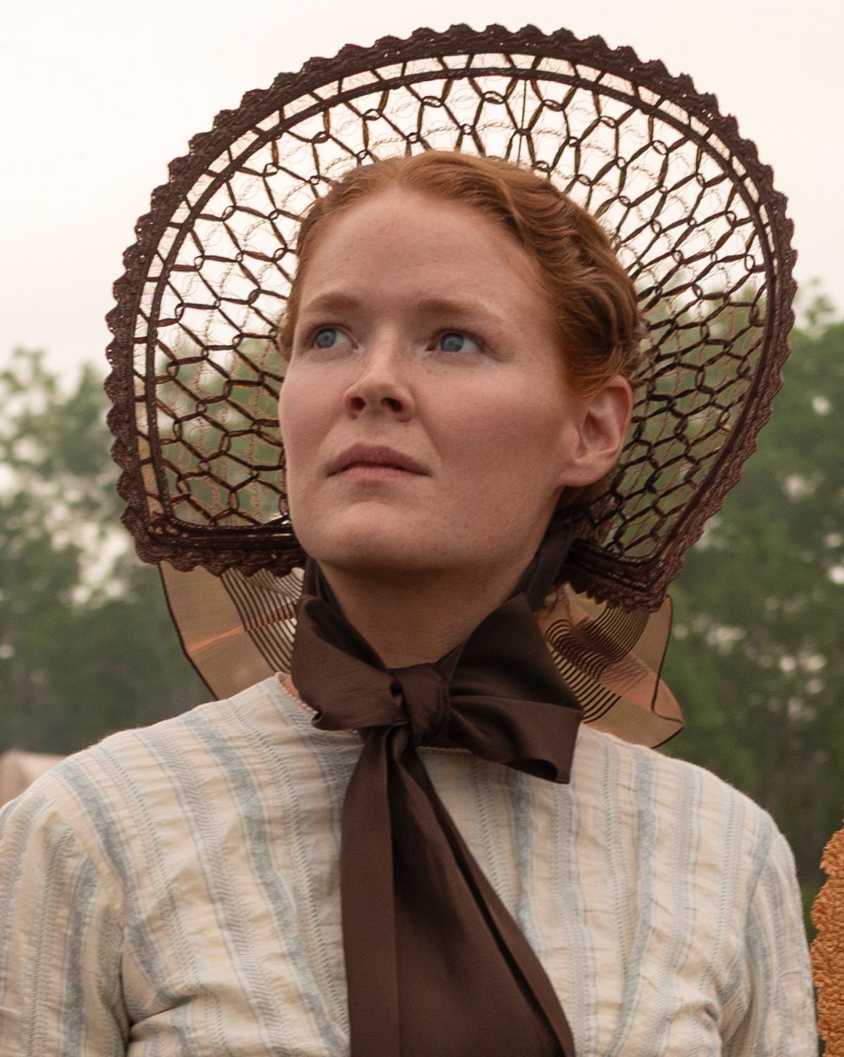 Woman in historical clothing and bonnet standing outdoors, looking upward with a serious expression, trees blurred in the background, overcast sky.