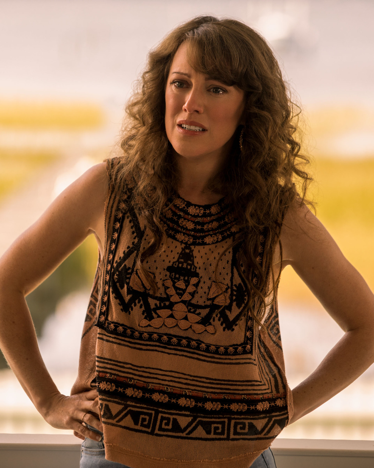 Samantha Soule as Anna Carrera with curly brown hair wearing a patterned sleeveless top stands indoors with hands on hips, looking concerned. Blurred outdoor scene with greenery and bright light in the background.