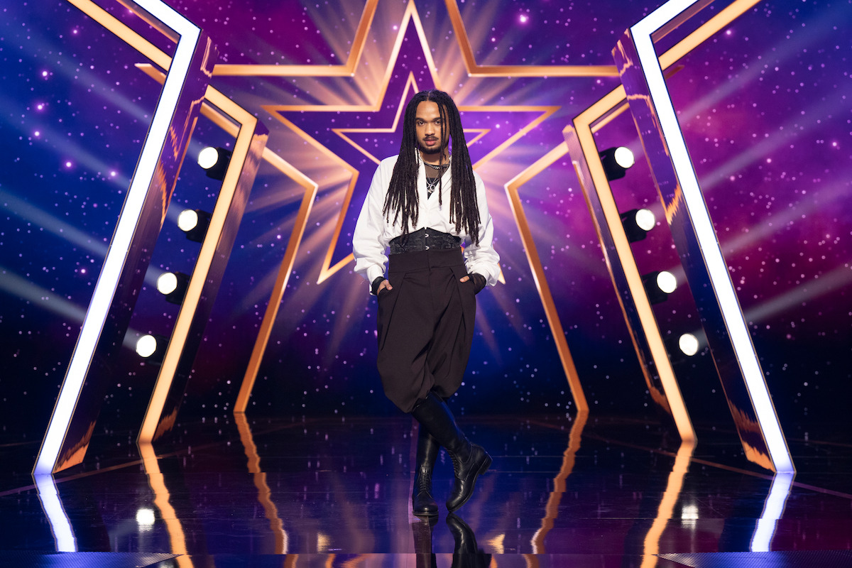 Jaïr Faria with long dreadlocks poses confidently on a shiny stage with dramatic lighting and a large illuminated star backdrop, surrounded by purple and gold lights.