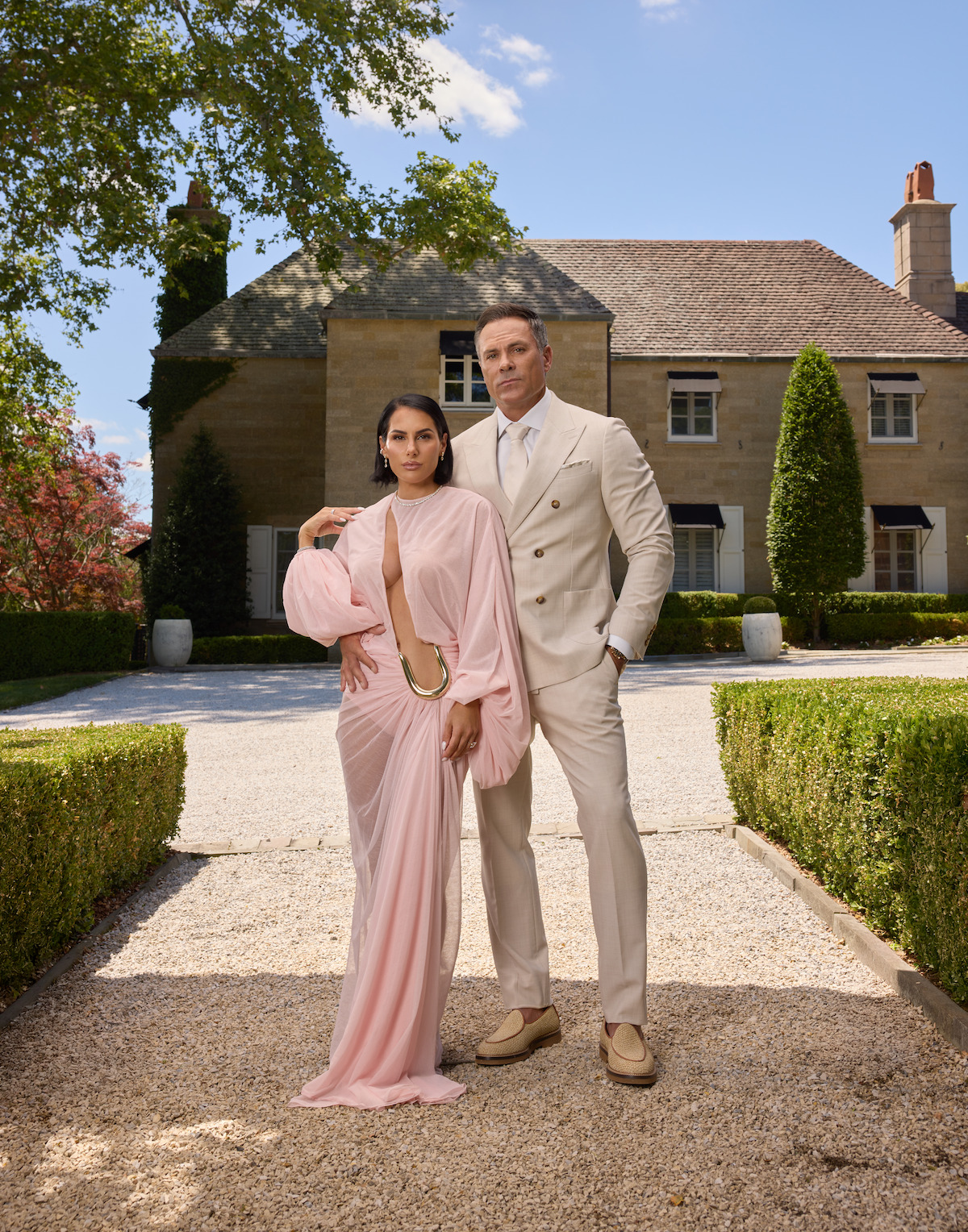 A well-dressed couple stands on a gravel path in front of a large, elegant house with manicured hedges and greenery on a sunny day. The woman wears a pink gown and the man wears a beige suit.