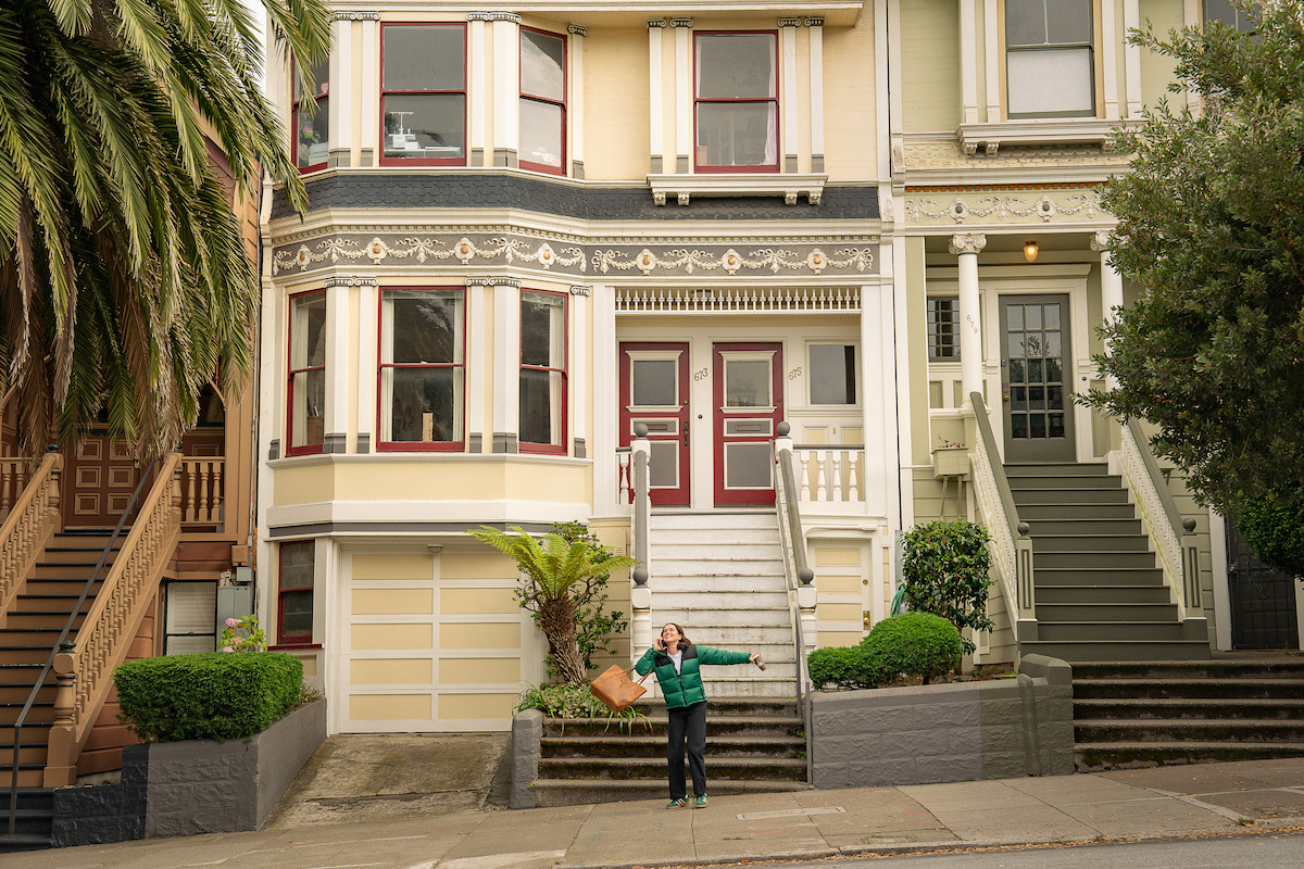 Person standing near the steps of a classic Victorian-style house in a residential neighborhood, holding a bag, with greenery and trees around the entrance on a cloudy day.