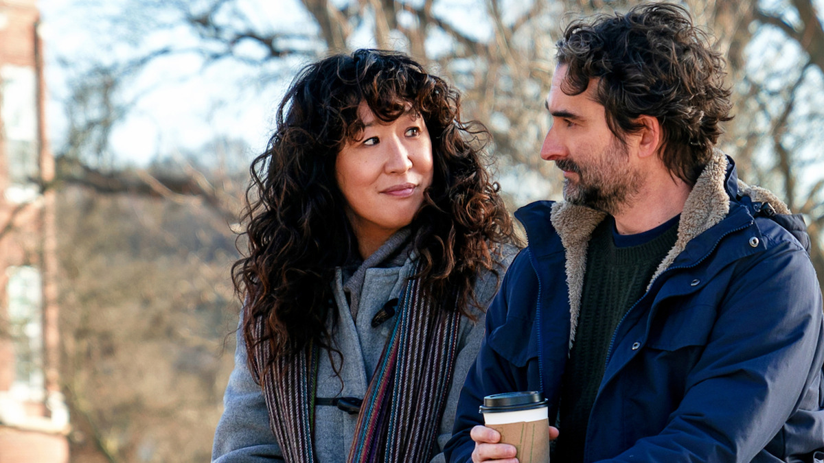 Two people standing outdoors in winter coats, looking at each other and smiling. One is holding a coffee cup. Background includes bare trees and a building, suggesting a chilly day in a park or campus setting.