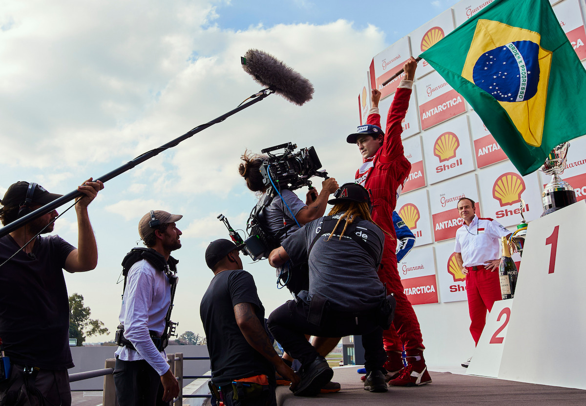 Behind the scenes with Gabriel Leone as Ayrton Senna. A camera crew crouch below the actor as he raises his arms and a Brazilian flag in the air. Go Senna!