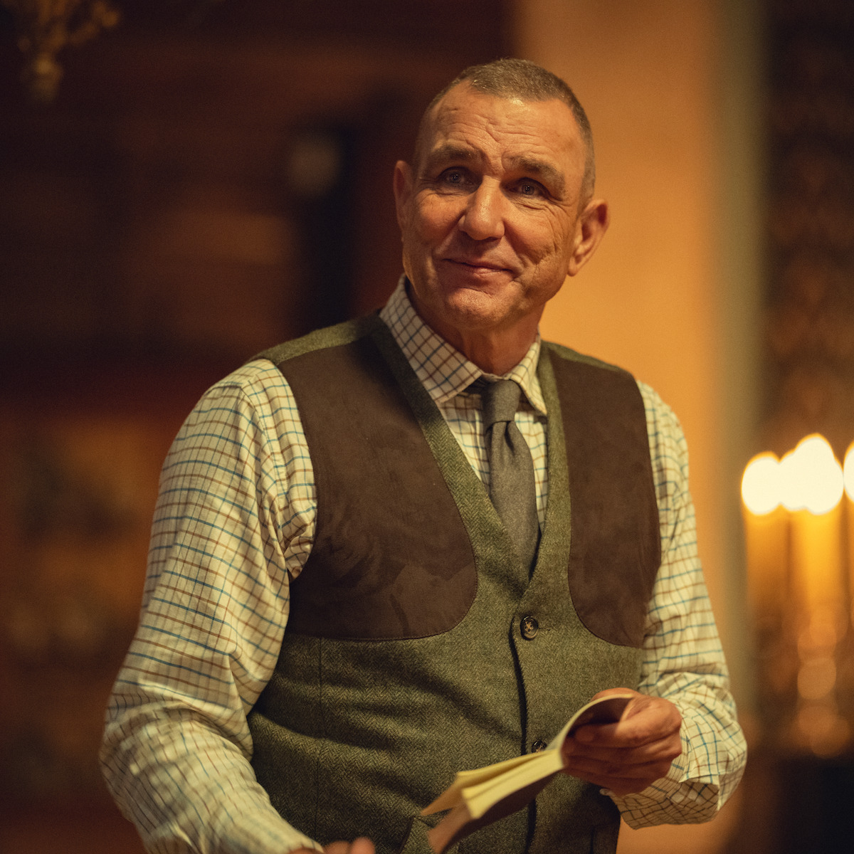 A middle-aged man in a checkered shirt, vest, and tie is holding an open book and smiling in a warmly lit, indoor setting with candles in the background.