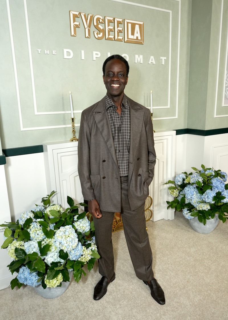 Man in a brown suit smiling and posing indoors at a formal event, with floral arrangements and decorative candles, standing in front of a wall with “FYSEE LA” and “The Diplomat” signage.