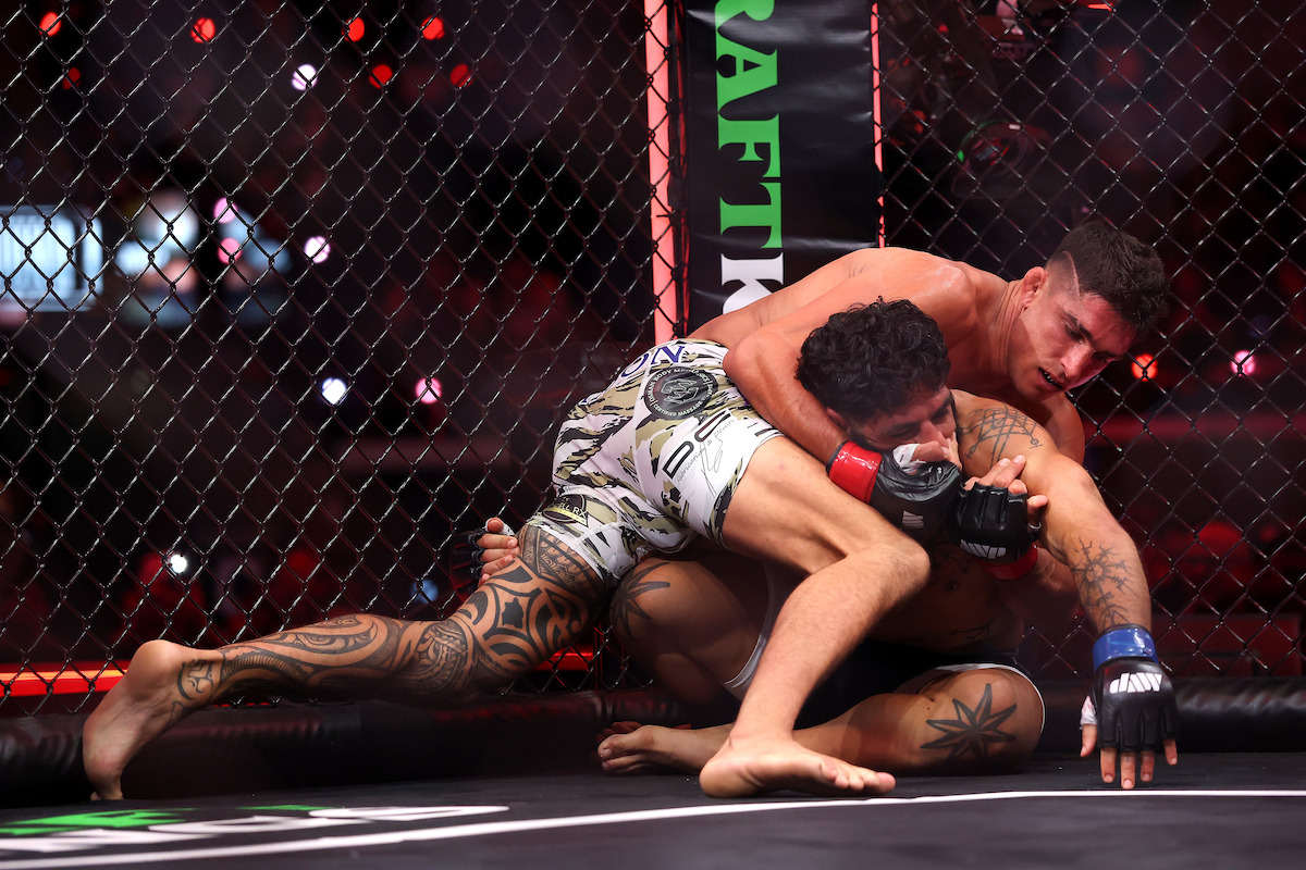 Two mixed martial artists with tattoos are grappling on the ground inside a caged octagon, with one fighter attempting a choke hold near the edge of the mat during a professional MMA match.