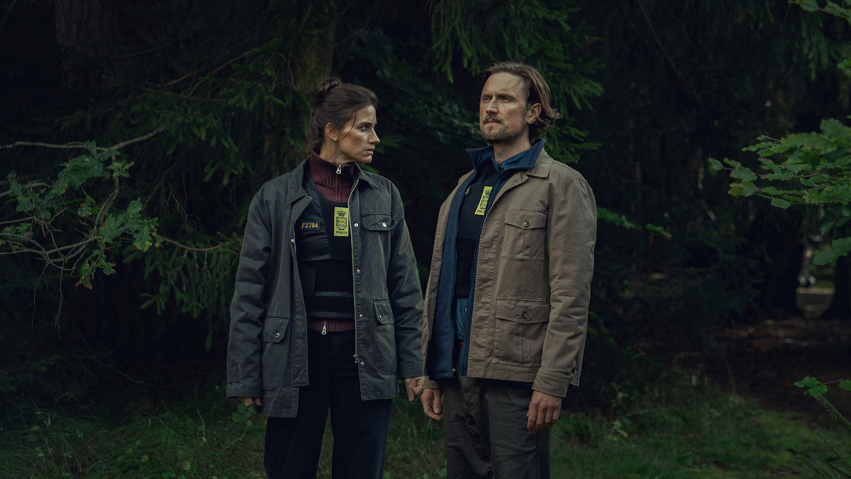 Two people wearing badges and jackets stand side by side in a dense, green forest, holding hands and looking in opposite directions, suggesting a serious or investigative scenario in a natural outdoor setting.