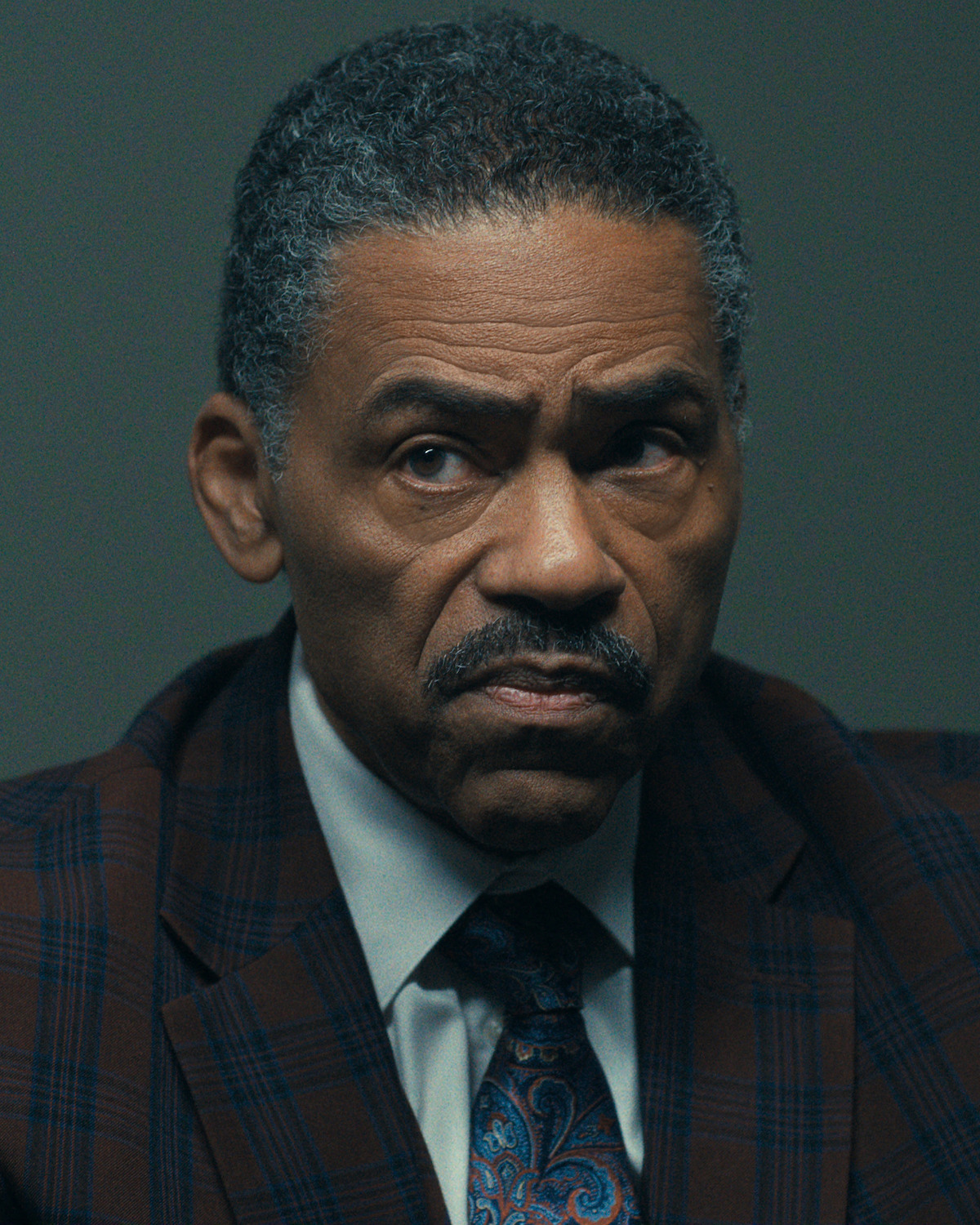 Middle-aged man in a plaid suit jacket, dress shirt, and paisley tie sits indoors under soft lighting, appearing serious and focused against a plain dark background.