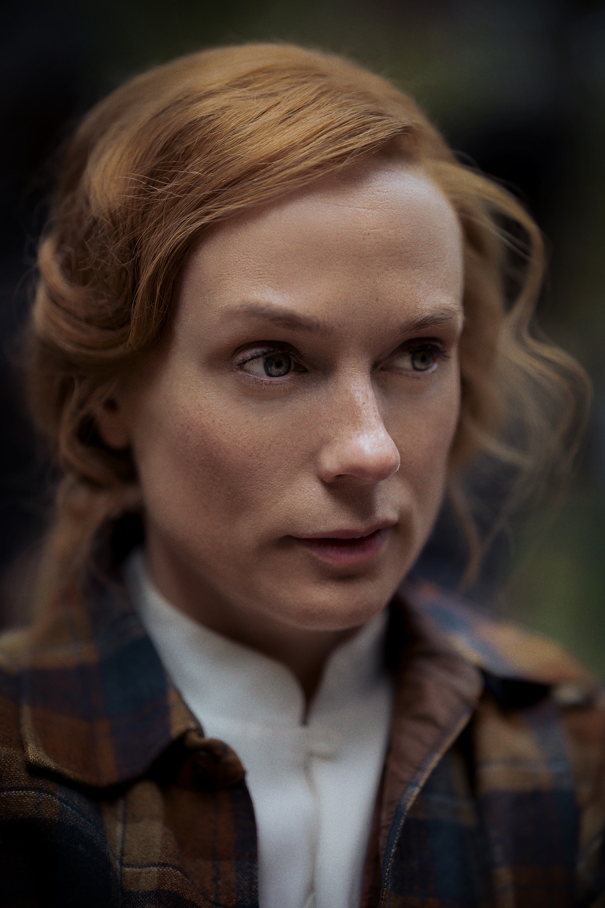 A woman with red hair wearing a plaid jacket and white shirt stands outdoors, looking thoughtful. The background is blurred, creating a focused and contemplative mood.