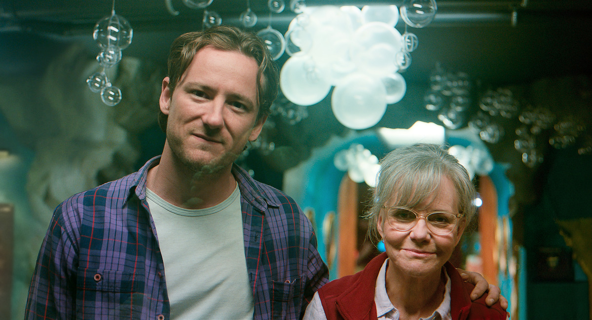 Lewis Pullman as Cameron and Sally Field as Tova stand together indoors in a colorfully decorated room with artistic bubble-like light fixtures and blue-toned walls, both smiling gently at the camera.