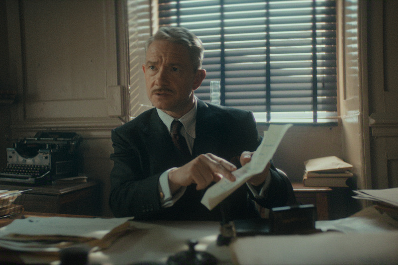 Man in a suit sits at a cluttered desk in a dimly lit office with venetian blinds, holding and reviewing paperwork, conveying a serious and focused mood. Old-fashioned decor and typewriter suggest a vintage setting.
