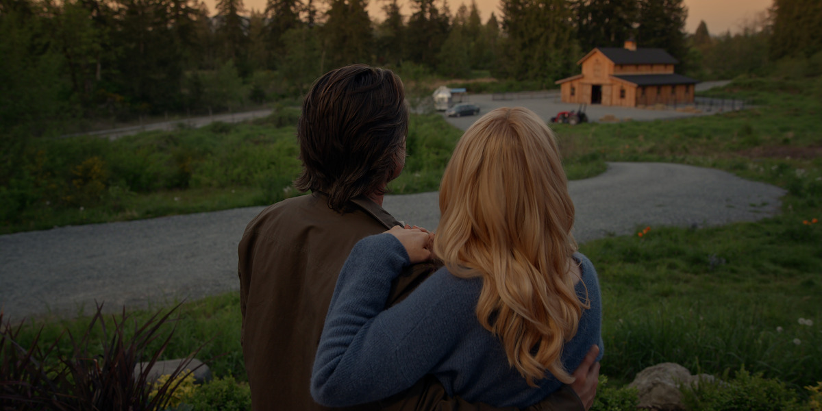 A couple stands closely together, looking at a wooden house in a rural, green landscape at sunset, with trees and a winding driveway in the background, creating a peaceful, intimate setting.