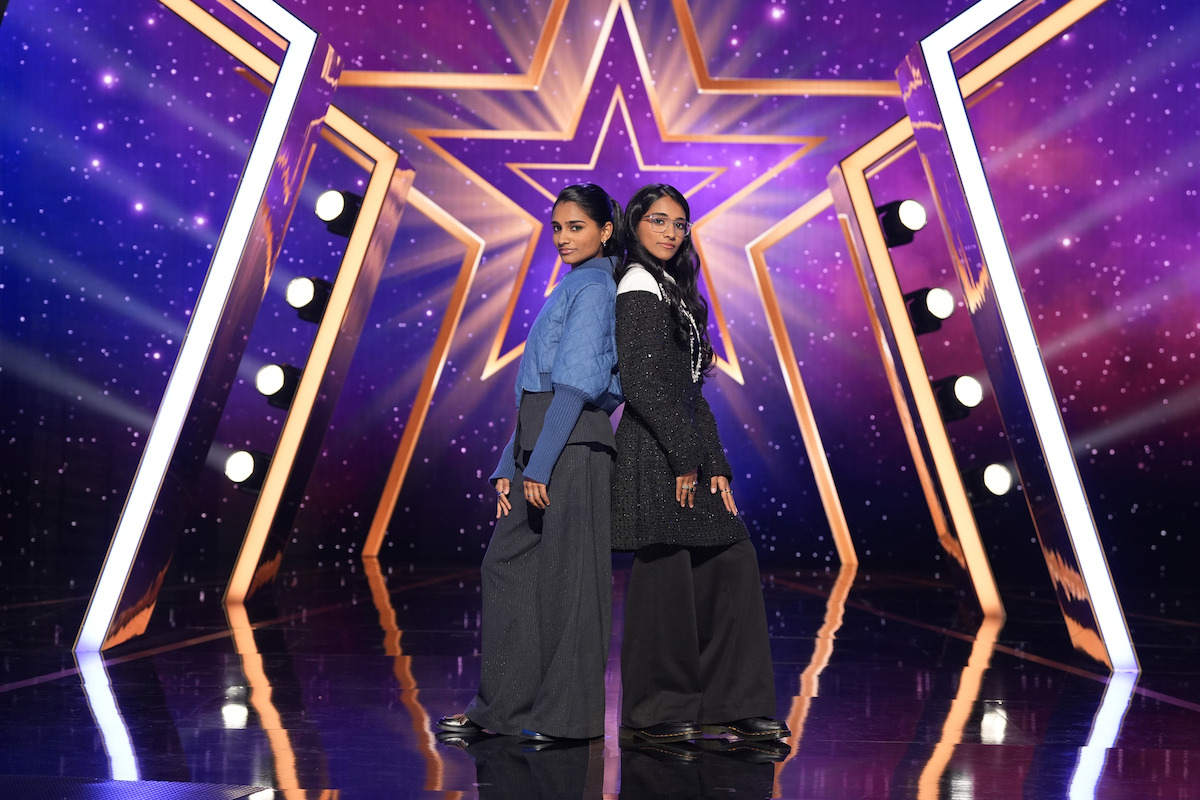 Kiran and Nivi stand back to back on a shiny stage with dramatic lighting, a large illuminated star, and purple cosmic-themed background, creating a glamorous talent show or entertainment event atmosphere.
