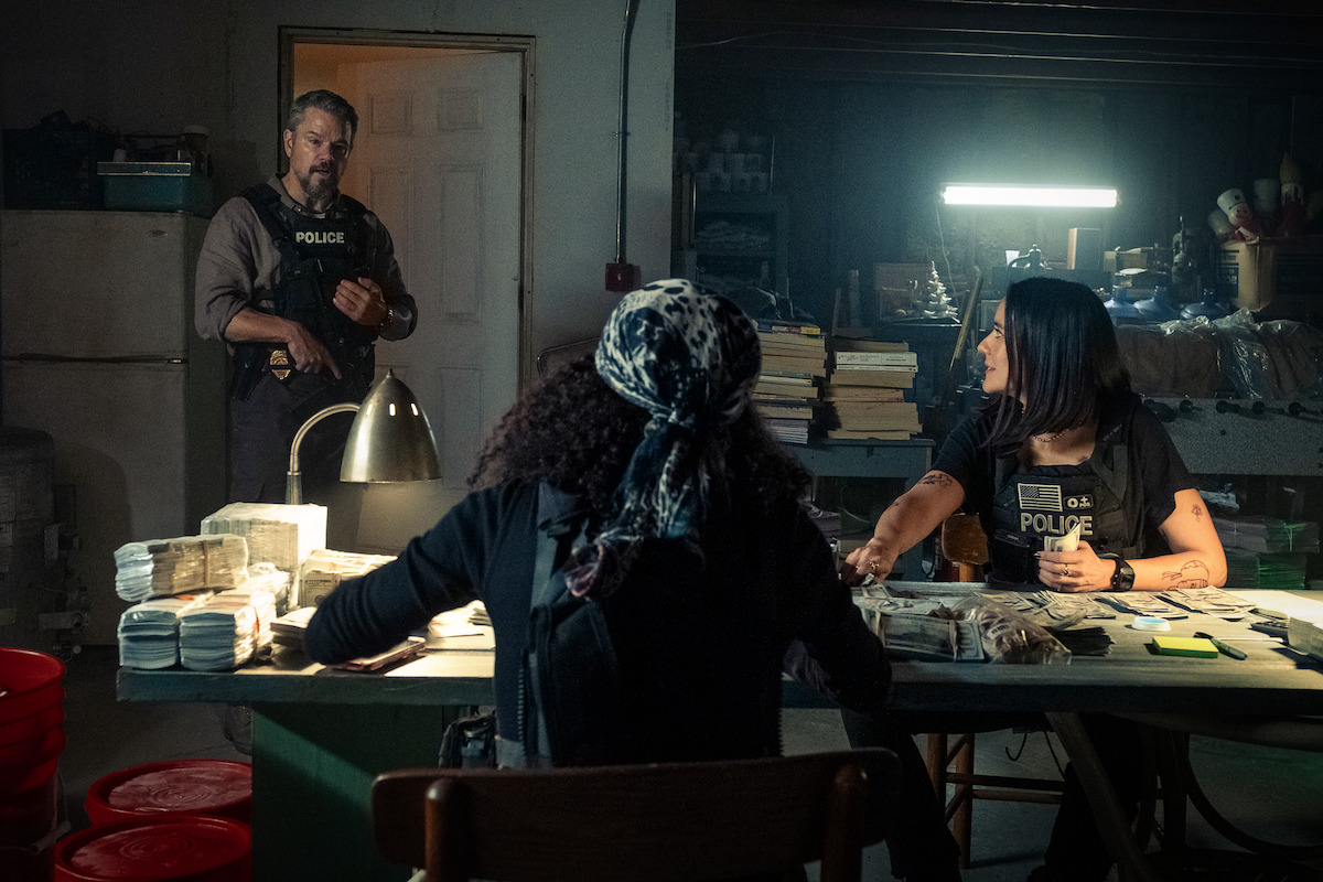 Three police officers in a dimly lit room surrounded by stacks of money on a table, industrial shelves, and evidence bags, suggesting a crime investigation or drug bust setting.