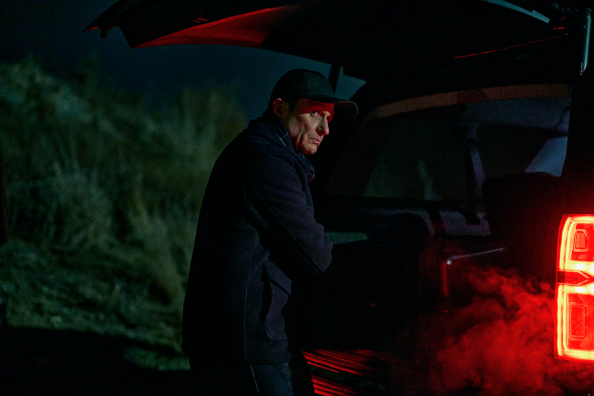 A man in a dark jacket and cap stands by the open trunk of an SUV at night, illuminated by red tail lights and surrounded by a mysterious outdoor setting, creating a tense, suspenseful mood.