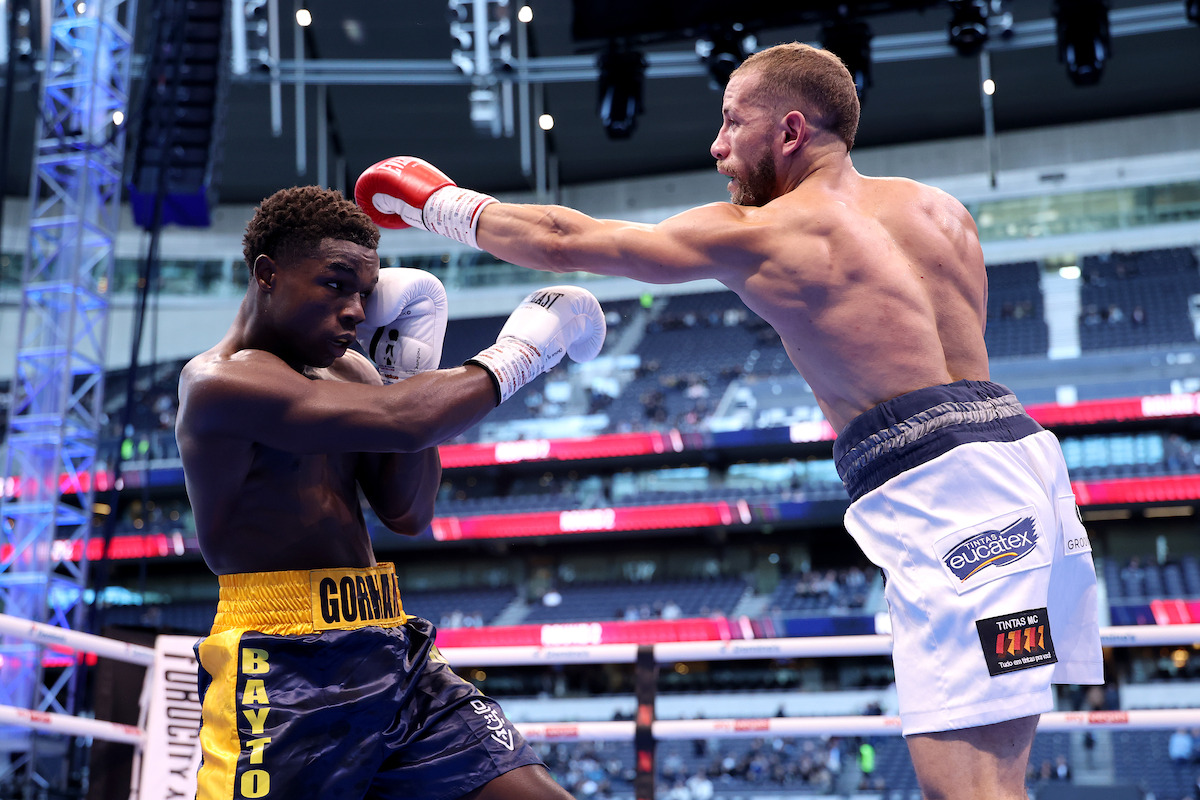 Two male boxers in a boxing ring during a match; one boxer throws a punch while the other defends, with stadium seating and spectators in the background, under bright sports lighting.