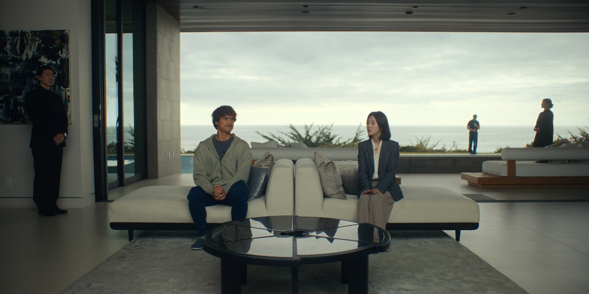 Two people sit apart on a modern white sofa in a spacious, stylish living room with large windows overlooking the ocean, while three others stand in the background both inside and outside near the sea.