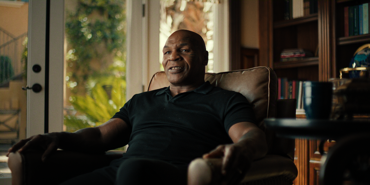 A man sitting in a cozy armchair in a warmly lit home, surrounded by bookshelves and large windows with sunlight and plants visible outside, a coffee table with a mug in the foreground.