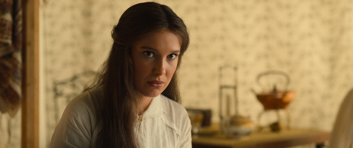 Young woman with long brown hair sits indoors, wearing a light blouse. The background shows a softly lit vintage room with floral wallpaper and a table holding a fruit basket and teapot, giving a warm, old-fashioned atmosphere.