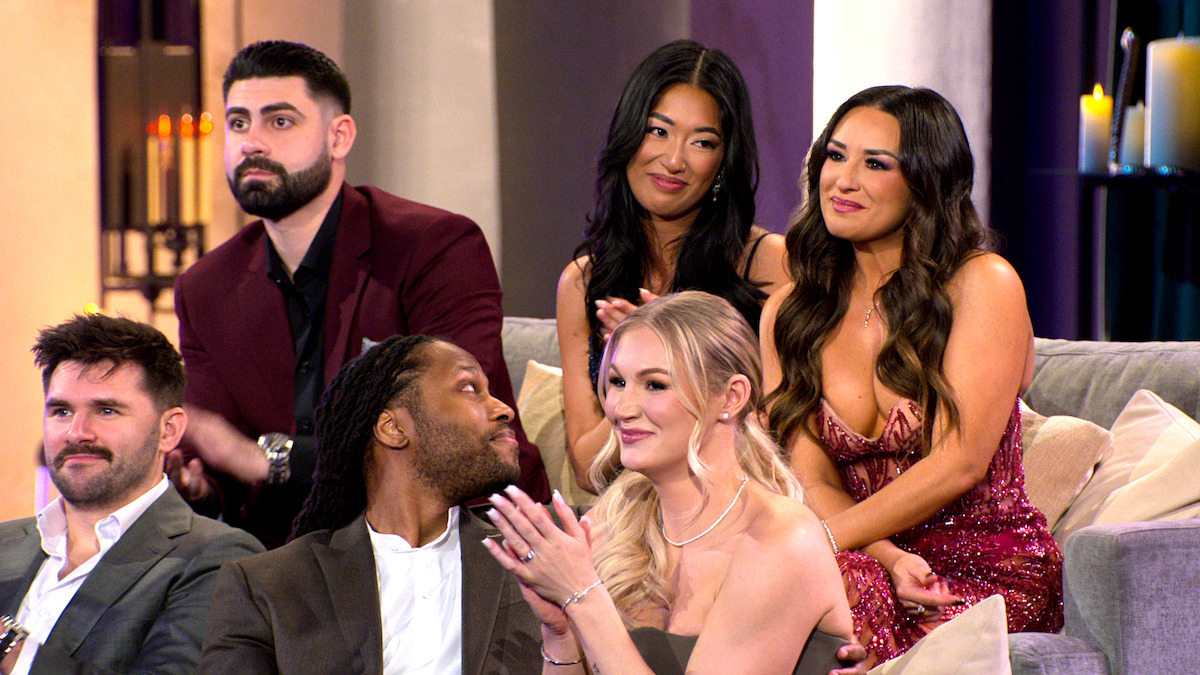Six people dressed formally sit on a couch and chairs, smiling and interacting in a warmly lit indoor setting with candles in the background, suggesting a social or reunion event.