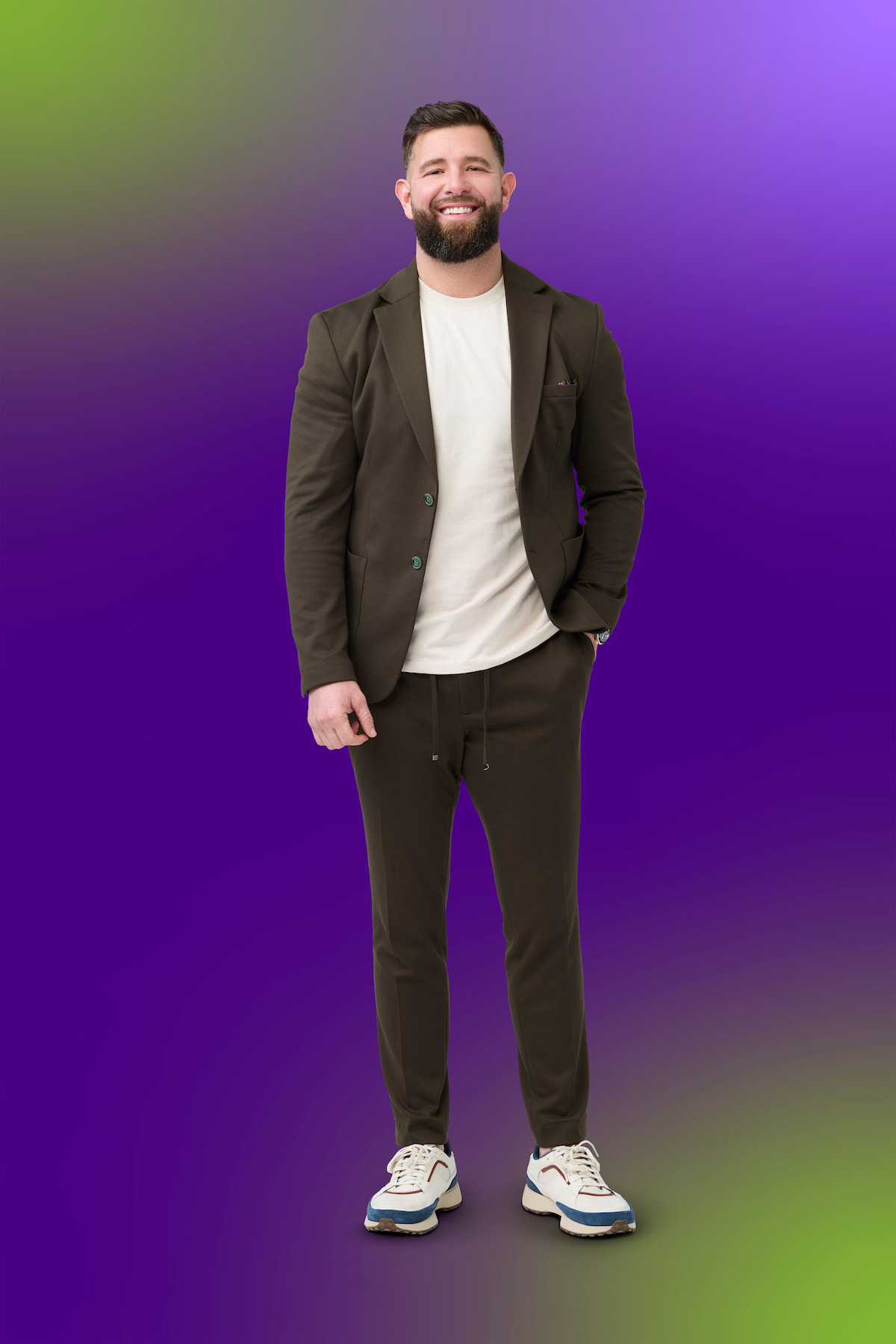 A bearded man in a dark casual suit with white sneakers and a white t-shirt stands smiling against a vibrant gradient background of purple, green, and yellow tones.