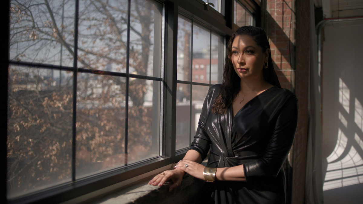 Woman in black dress stands by large sunlit window, indoors, with bare trees and buildings visible outside. Natural light casts shadows, creating a calm and contemplative atmosphere in a modern, loft-like space.