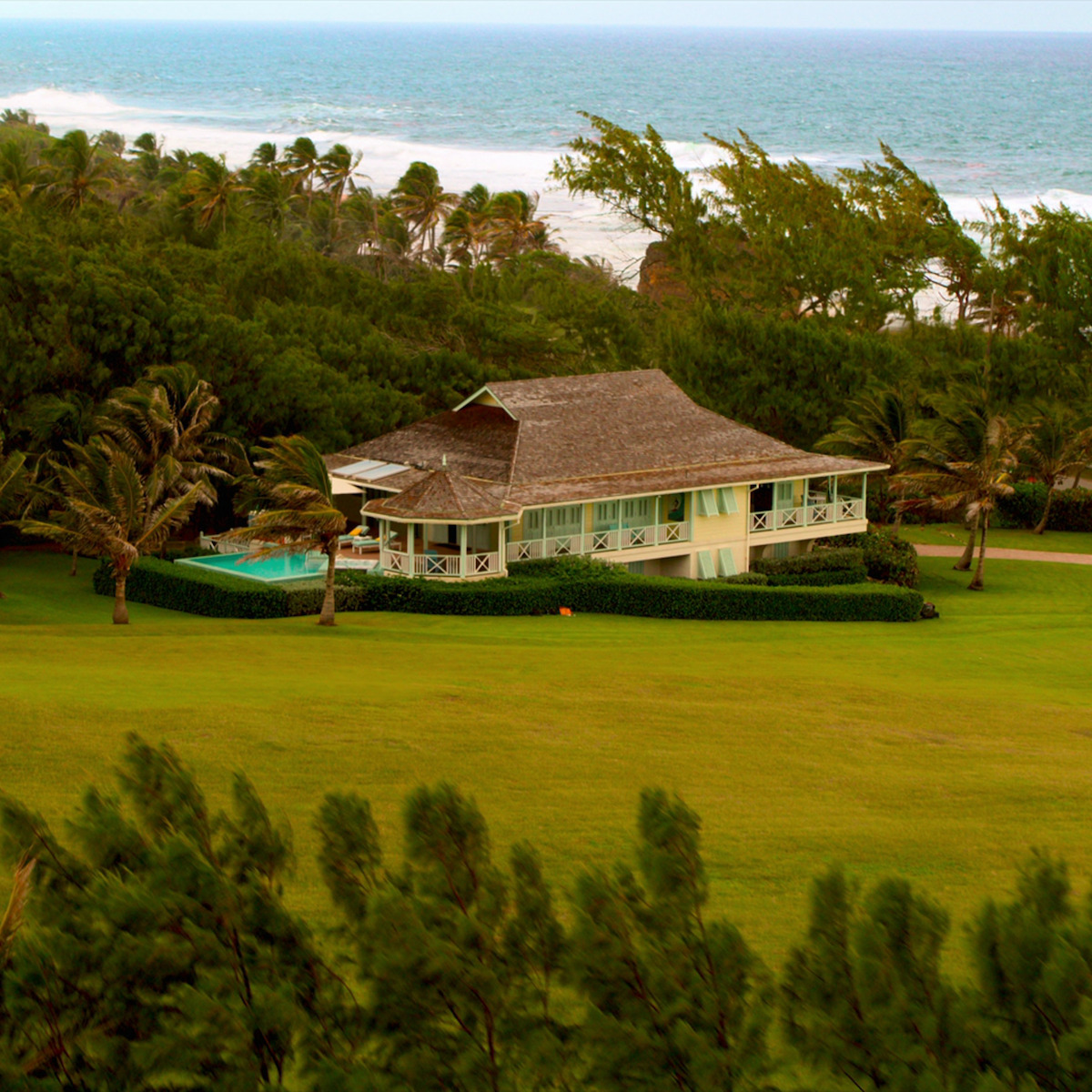 Uma casa na beira da praia com teto de palha cercada de árvores e uma paisagem verde exuberante, com piscina e o mar visto no fundo em um dia de vento.