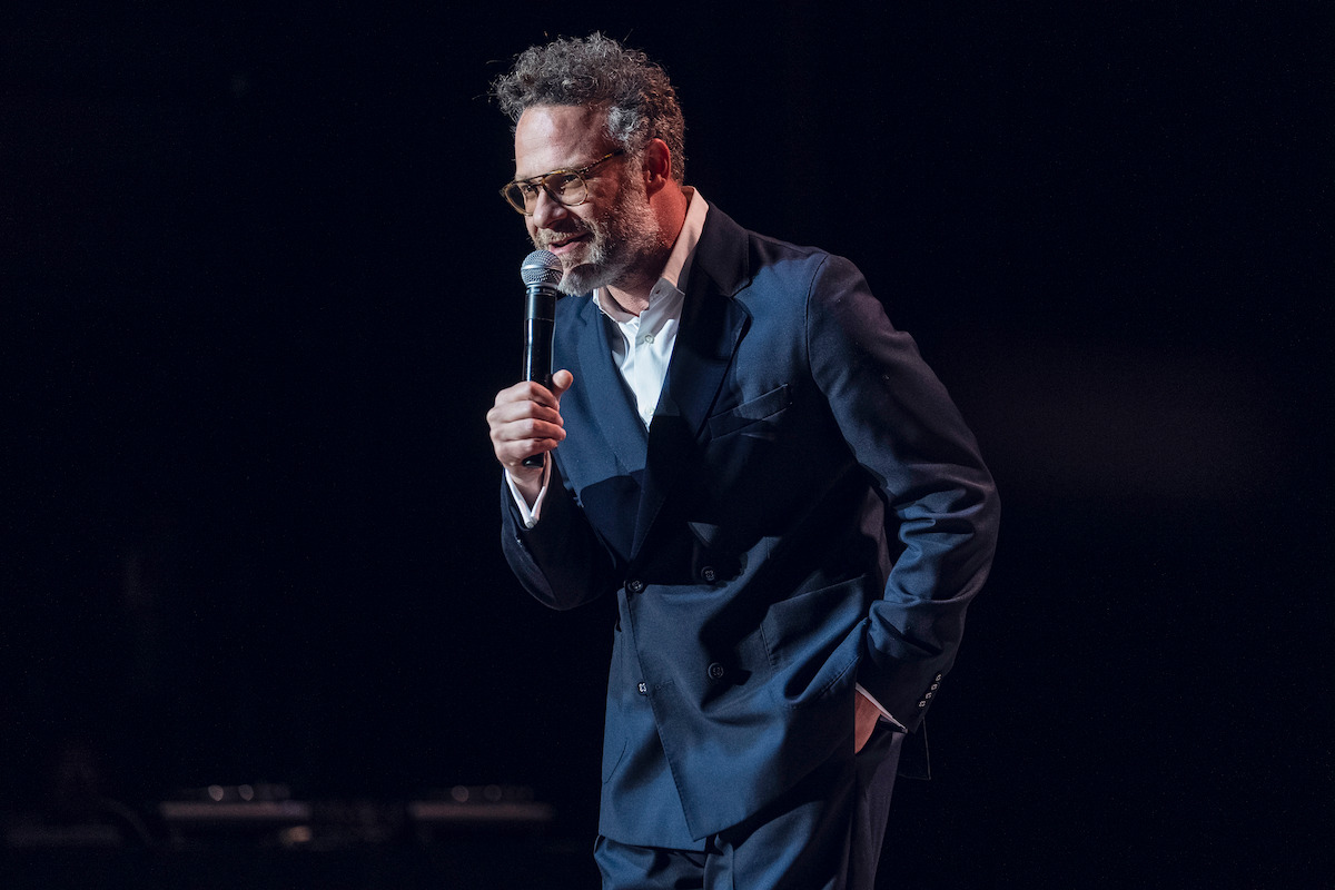 A man in a dark suit and glasses performs stand-up comedy on stage, holding a microphone, with a dark background and stage lights highlighting him.