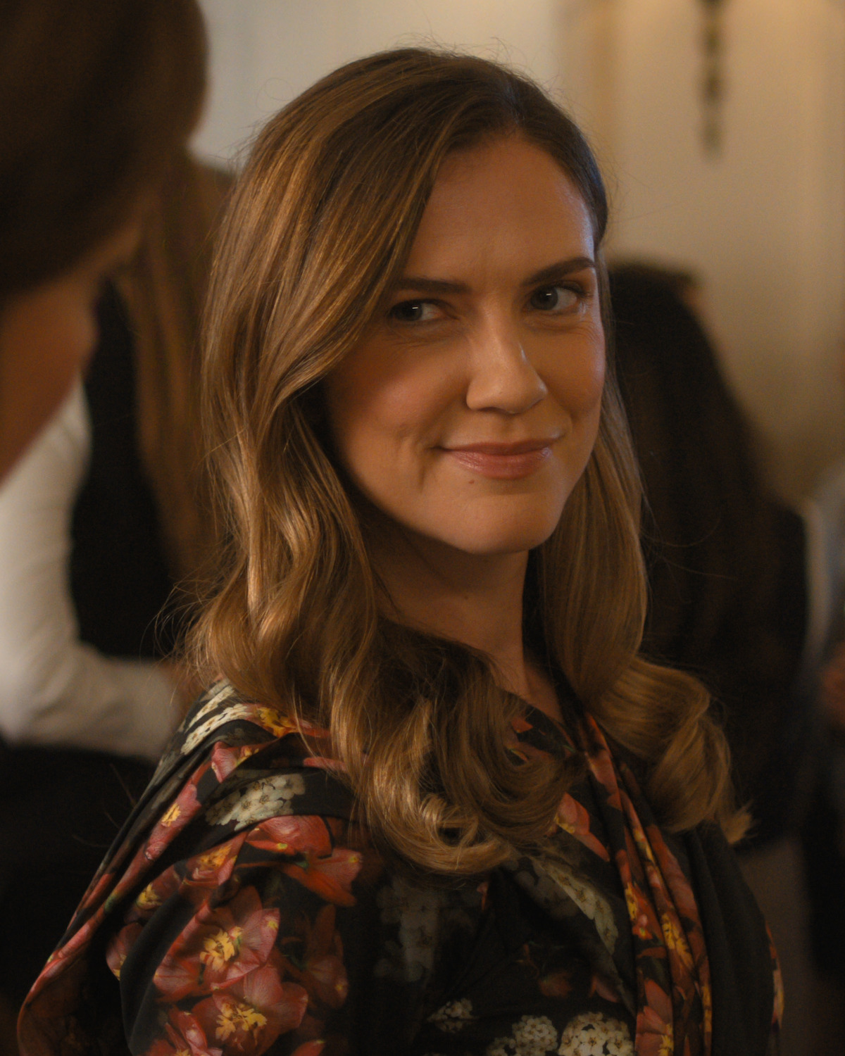 Woman with long, wavy brown hair in a floral dress smiling and looking at the camera in an indoor setting with other people blurred in the background, suggesting a social or formal gathering.