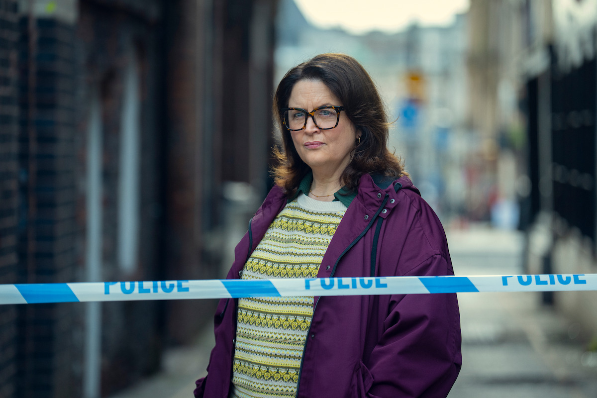 A woman wearing glasses and a purple jacket stands behind police tape on a city street, suggesting a crime scene. The background is blurred, the mood appears serious and investigative.