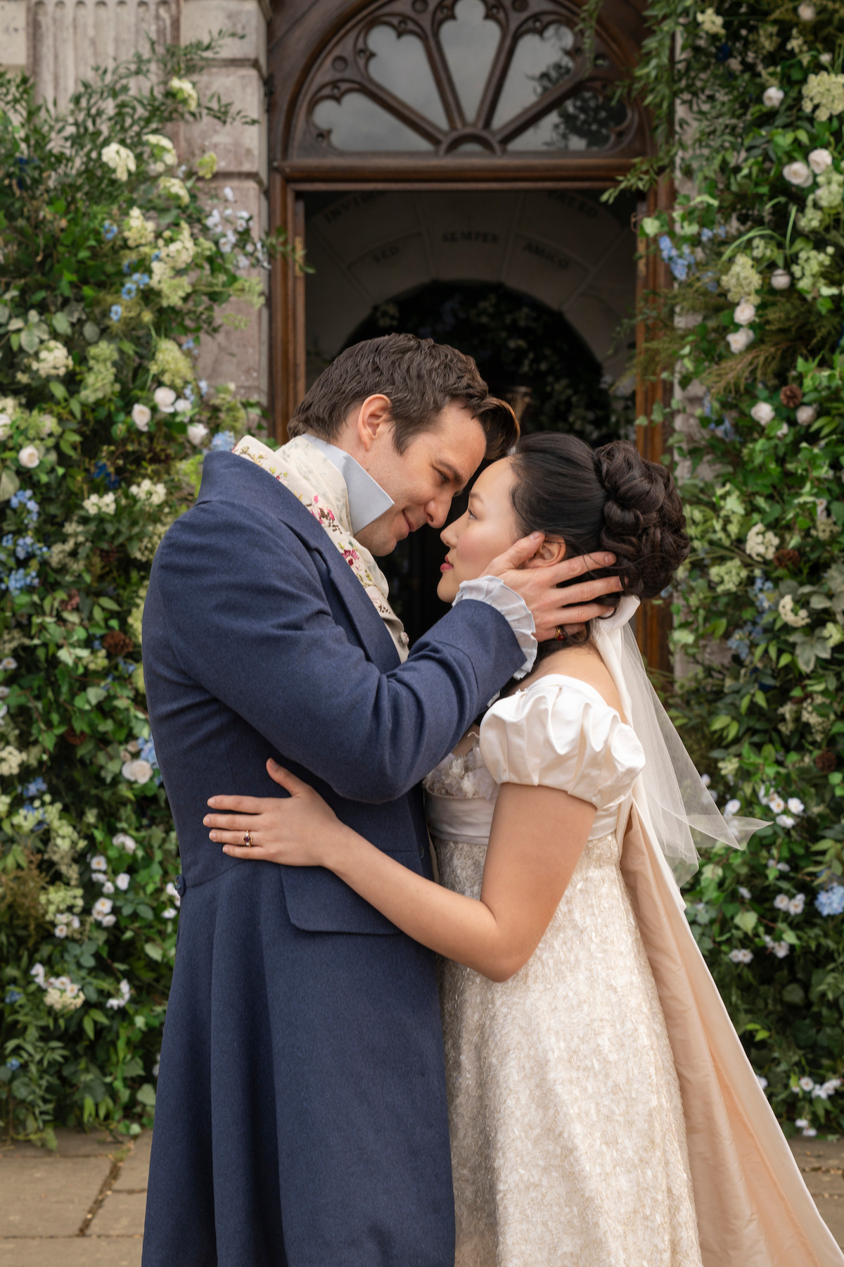 A couple in historical attire embraces and touches foreheads outside a flower-adorned archway, suggesting a romantic or wedding scene in a lush, elegant garden setting.