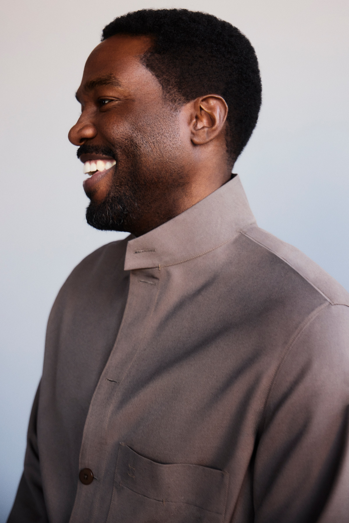 A man with short black hair and a beard smiles while looking to the left, wearing a high-collared taupe jacket in front of a plain, light-colored background.