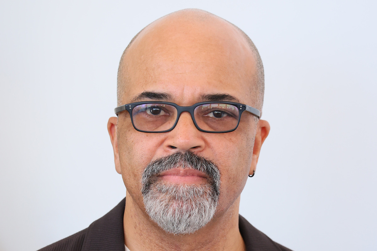 Bald man with glasses, grey goatee, and brown jacket looking into the camera, neutral expression, plain light background, calm and professional atmosphere.