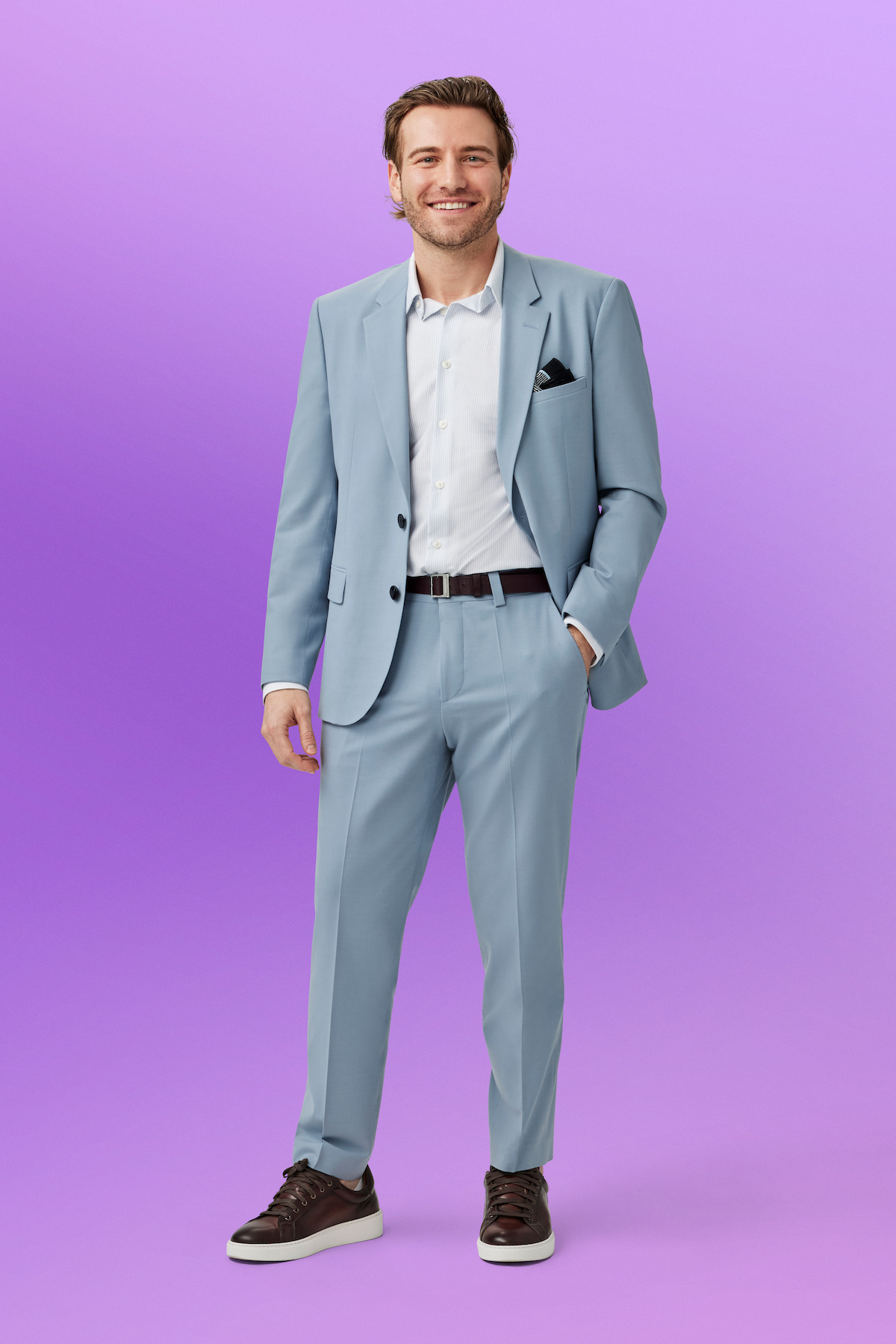 Steven standing and smiling in a light blue suit with white shirt and black shoes, against a solid purple background.