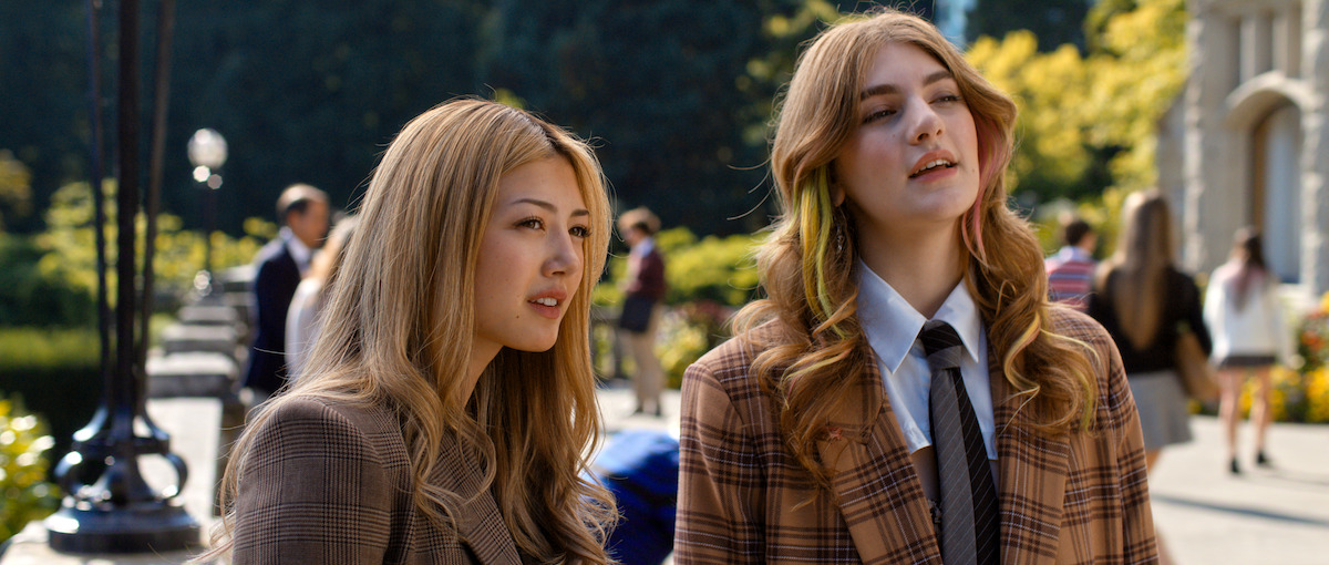 Two young women in brown plaid jackets stand outdoors on a sunny day in a city park or campus, surrounded by people, greenery, and buildings, engaged in conversation.